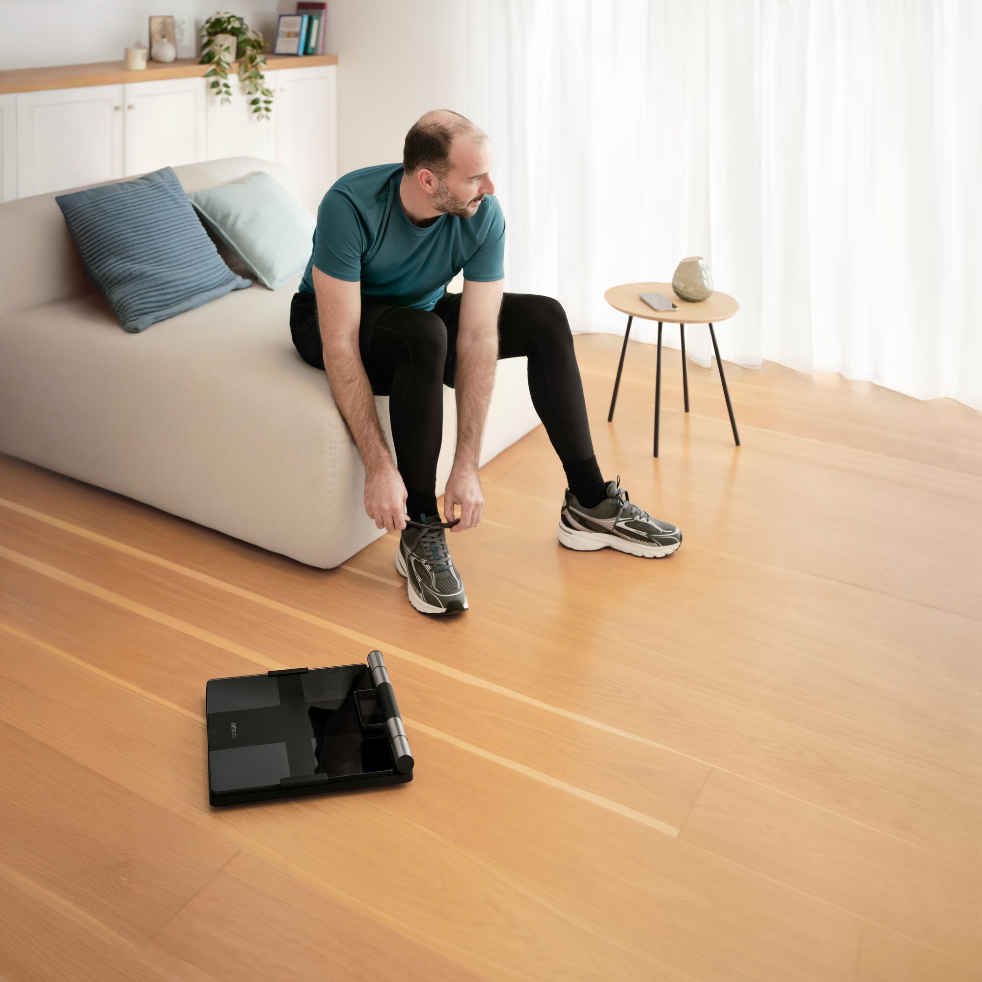 A man in workout clothing sits on a sofa tying his athletic shoe in a bright living room. An OMRON body composition monitor with attached handgrip lies on the wooden floor in front of him, ready for use.