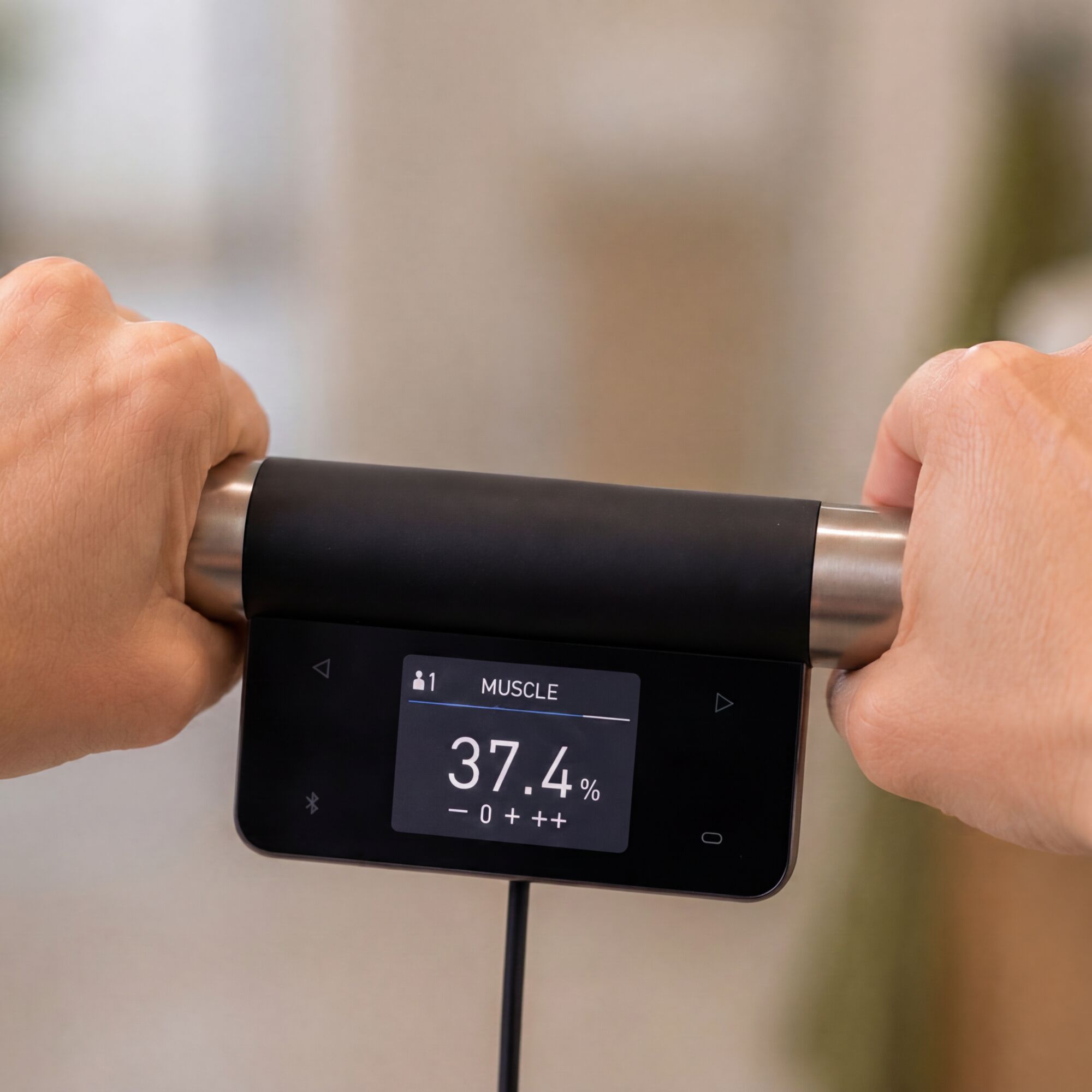 Close-up of two hands holding the metallic ends of an OMRON body composition monitor handgrip. The central digital display shows the Muscle measurement for user 1 as 37.4%, with a scale labeled minus, 0, plus, and double plus beneath the result.
